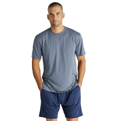man facing front with hands in pockets wearing a grey short sleeve shirt and blue shorts